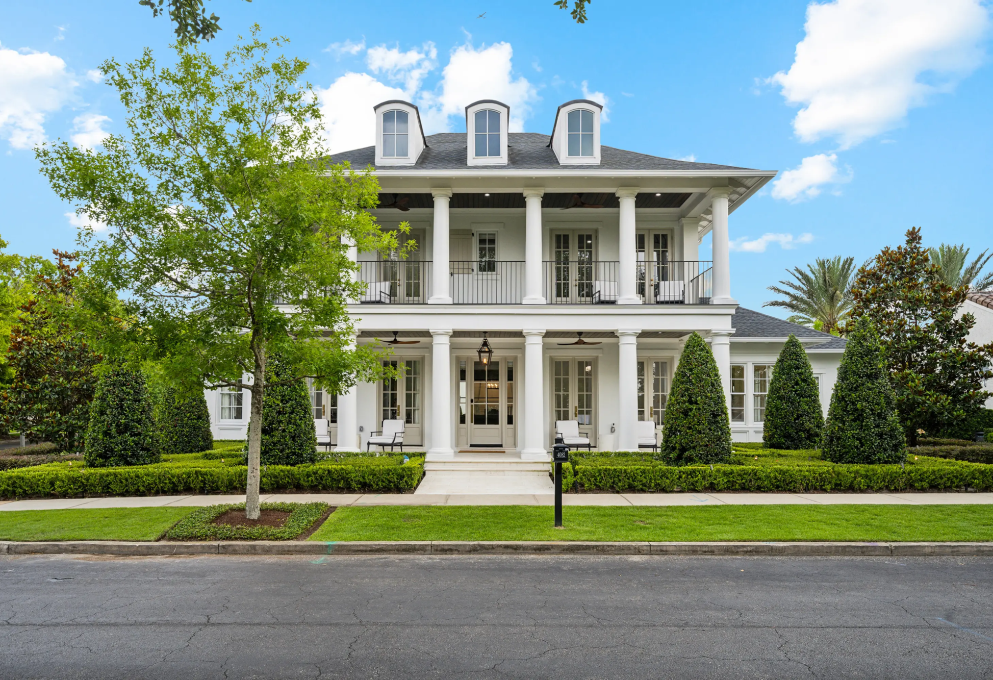 Two-story white Southern colonial home with double-tier columned porches, framed by a manicured boxwood hedge border and precisely shaped conical holly topiaries flanking the front walkway.