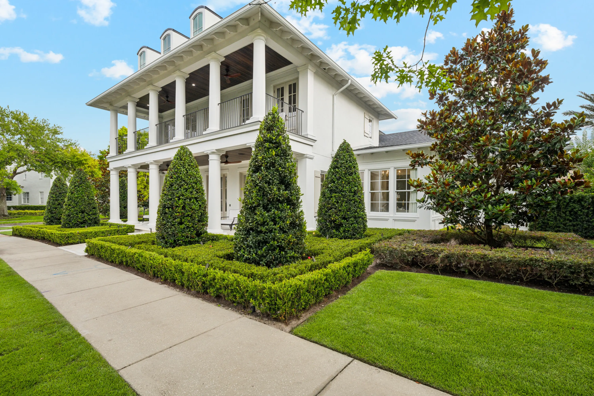 Side view of a white two-story home showcasing a row of tightly pruned conical holly topiaries set within a crisp boxwood hedge border along the front lawn.