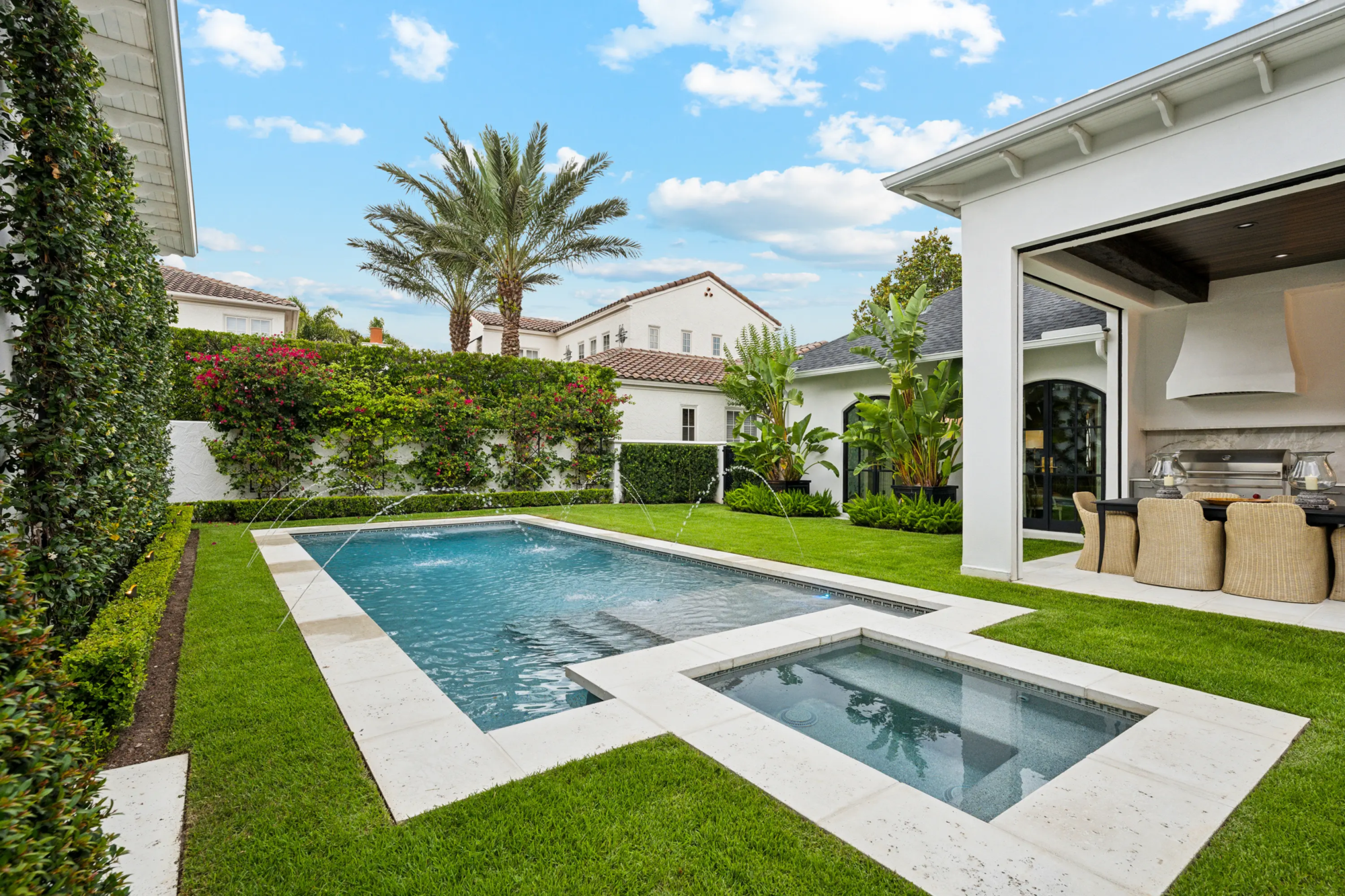 Full courtyard view of pool, spa, and outdoor kitchen area bordered by boxwood hedges, jasmine trellis wall, bougainvillea hedge, and potted bird-of-paradise plantings.